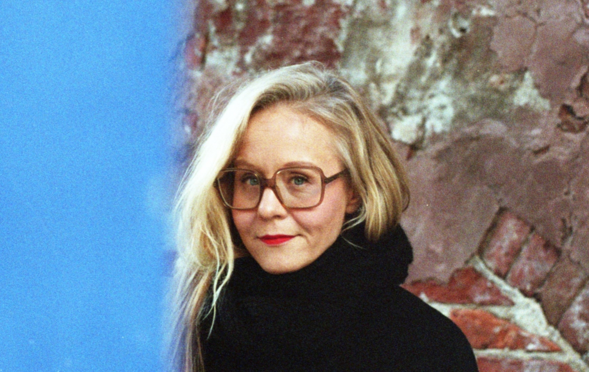 Anna-Kaari Hakkarainen katsoo kameraan. Seisoo punaista tiiliseinää vasten.
